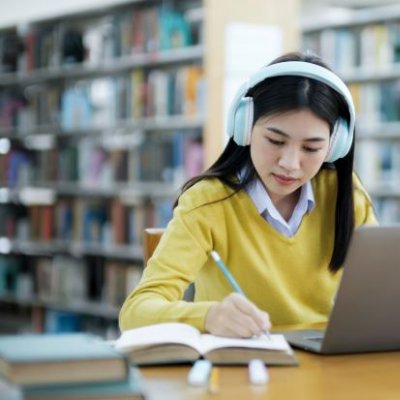 a person with black hair wearing a yellow top and pale blue headphones sits at a desk with a laptop and open books